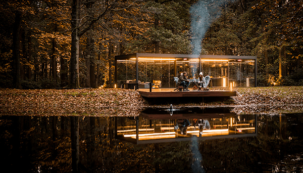 Opplyst pergola i glass ved et vann i høstskog, med folk rundt bordet